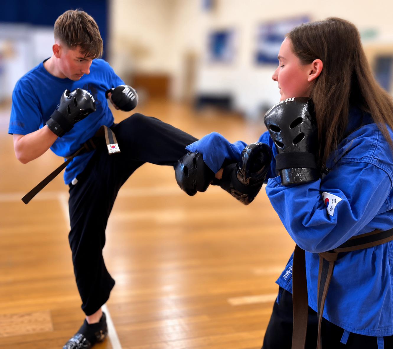 Two teens in Choi Kwang Do sparring gear, sparring with pads on a wooden floor